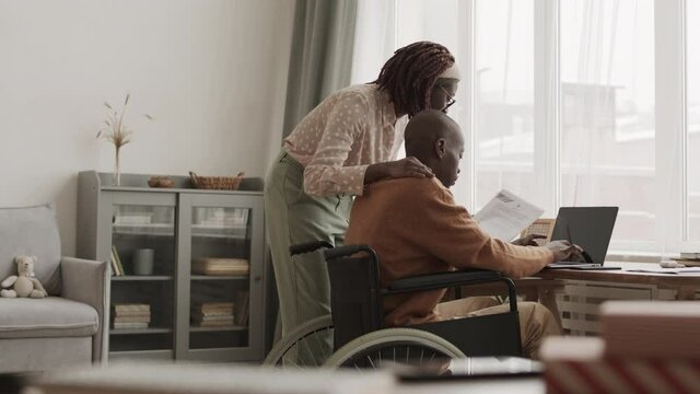 Medium shot of young African woman standing at desk with document in hand while young African disabled man sitting on wheelchair and typing on laptop keyboard
