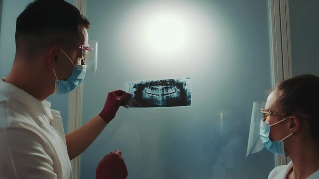 Radiologist and the assistant physician examine the panoramic x-ray of the person's jaw and discuss the complexity of the treatment. The doctor points to problem teeth on an X-ray