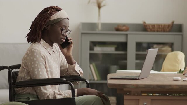 Zoom out of young attractive African disabled woman wearing casual clothes sitting on wheelchair at desk in living room and having business telephone conversation