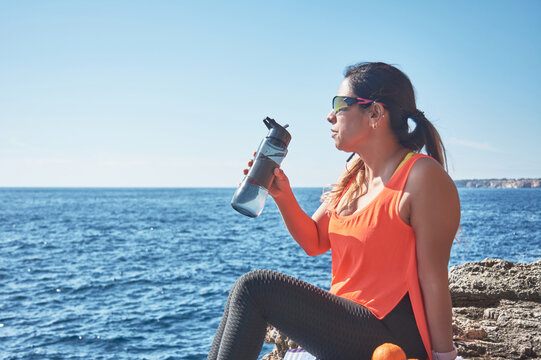 Latin Woman, Middle-aged, Resting, Regaining Strength, Eating, Drinking Water, After A Gym Session, Burning Calories, Keeping Fit, Outdoors By The Sea, Wearing Headphones And Smart Watch