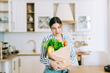 Young smiling caucasian woman hold eco shopping bag with fresh vegetables and baguette in modern kitchen.