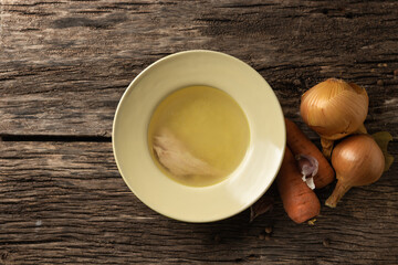Bowl with clear rich broth. White crockery on an aged wooden table. Vegetables - carrots, onions, garlic, spices