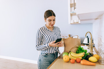 Young smiling caucasian woman use smartphone in the modern kitchen, bag with fresh vegetable on the table. Online buying food and grocery.