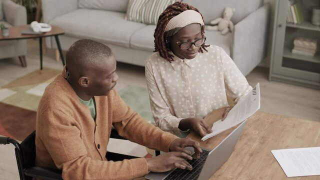 High angle view of young African woman sitting at desk with document in hands while young African disabled man sitting on wheelchair and typing on laptop keyboard