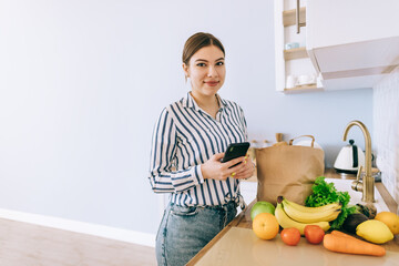 Young smiling caucasian woman use smartphone in the modern kitchen, bag with fresh vegetable on the table. Online buying food and grocery.