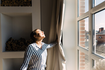 Young caucasian woman opens the curtains and looks out the window on a sunny morning