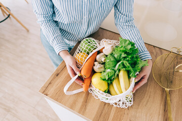 Caucasian woman hold eco net bag with fresh vegetables in modern kitchen.