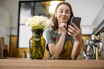 Young housewife in green apron ordering with smartphone a bouquet of fresh tulips. Florist with flowers on the kitchen at home.