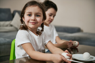 Adorable little girl sitting near a school boy, holding a paintbrush and smiling to camera before coloring Easter eggs.