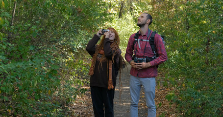 young couple of travelers with binoculars and photo camera walk in autumn forest	