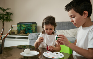 Boy and girl dressed alike coloring Easter eggs with paints sitting at table at home