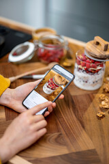 Digital recipe of healthy breakfast with granola on the phone screen on wooden table top in kitchen with ingredients on background. Phone in the hands.