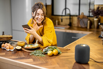Happy woman in bathrobe controlling home devices with a voice commands, speaking to a smart column...