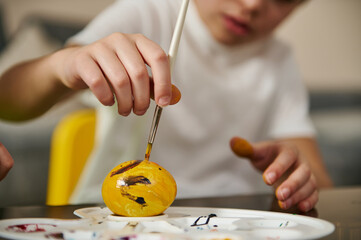 A yellow Easter egg on a palette and a boy's hand holding a paintbrush and decorating it for Easter.