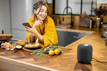 Happy woman in bathrobe controlling home devices with a voice commands, speaking to a smart column...
