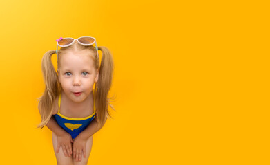 Little funny child in a blue swimsuit grimaces on a yellow background. A cute girl with ponytails on her head and sunglasses looks into the frame in surprise. Horizontal format. Place for text