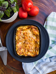 Delicious lasagna bolognese on a black ceramic plate, with cutlery, tomatoes, vase with basil and striped cloth on wooden base. Top view.