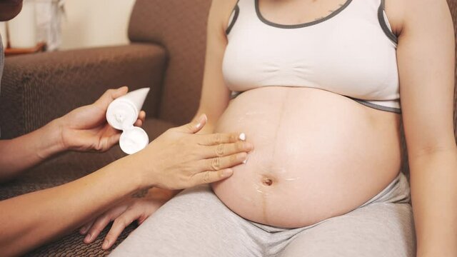 Asian Husband applying pregnancy cream to pregnant woman sitting on sofa at home, New family baby born concept.