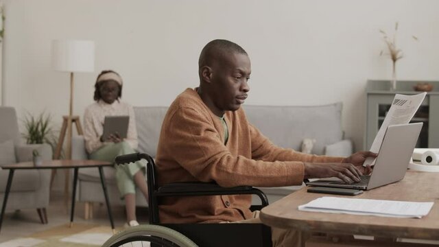 Lockdown of young African disabled businessman sitting on wheelchair at desk in living room, working with documents using laptop while his wife using tablet on background