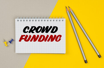 On a yellow-gray background are pencils, buttons and a notebook in which it is written - CROWD FUNDING