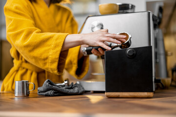 Woman in yellow bathrobe making espresso drink on a professional coffee machine at home, close-up. The process of making coffee on a carob machine.