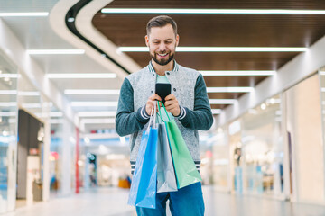 Attractive young casually dressed bearded man using his mobile phone while walking in the mall with a bunch of shopping bags in hand