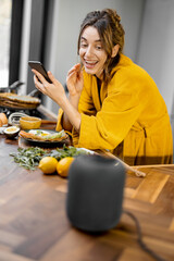 Happy woman in bathrobe controlling home devices with a voice commands, speaking to a smart column...