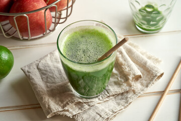 A glass of green juice, with paper straws, apples and lime in the background