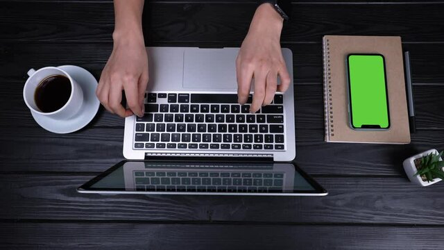 Top view of the hands of a girl who type, writes emails on an laptop. Near the laptop is a smartphone with a green screen and a chrome key. Place for your advertising content. Slow motion. Close up.