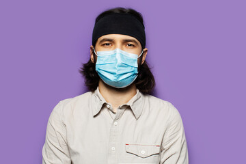 Studio portrait of young brunette guy, with long hair and protective medical face mask, on the background of purple wall.