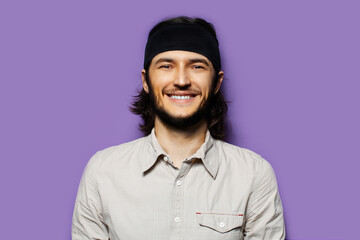 Studio portrait of young smiling man, with long dark hair and cheerful attitude, on the background of purple wall.