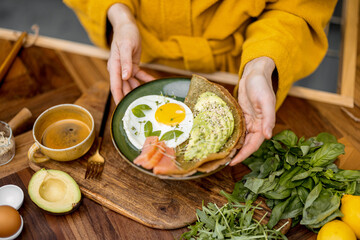 Healthy breakfast made of egg, sliced avocado, salted red fish and pancake on green plate on a wooden kitchen table, view from above