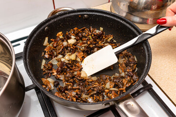 A woman is holding a silicone spoon and mixing fried mushrooms with onions in a frying pan on a gas stove.