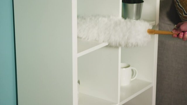 Young girl in a white sweater in a spacious bright room the color of a sea wave, with a window, wipes the dust from the lamp, beige sofa and white bookcase.