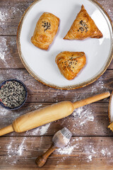 Chebureks on a plate, sauce, flour, sesame seeds, rolling pin on a brown wooden background.