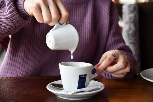 Young woman hands hold white ceramic milk jug with dripping drop into coffee cup. Pouring milk into crockery mug of warm drink with logo of Lavazza brand of italian coffee. Odessa, Ukraine, 01 20 2021