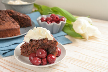 Juicy homemade chocolate cake with morello cherries and whipped cream, some tulip flowers and a blue napkin on a bright wooden table for a festive afternoon, copy space, selected focus