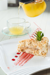 Napoleon cake decorated with a spiral of red chocolate, mint and berry jam on a white plate against a background of orange tea in a teapot and a transparent cup. Layered cake with pastry cream.