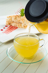 A waiter pours orange tea from a teapot into a transparent cup against the background of a napoleon cake decorated with mint and berry jam on a white wooden table