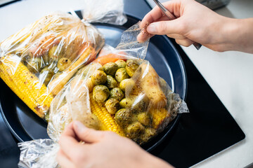 Male hands opening plant based baked dish with brussels sprouts, parsley root, corn. Mixed vegetables in a baking bag on the tray. Cooking healthy vegan lunch. Vegetarian cristmas dinner. Selective.