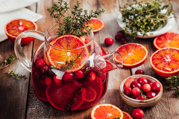 Citrus and berry tea with cranberries, blood oranges and thyme in a glass teapot on wooden table.