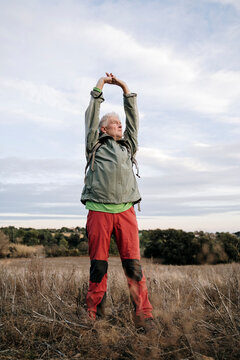 Senior male hiker stretching arms while standing on agricultural field against cloudy sky at countryside