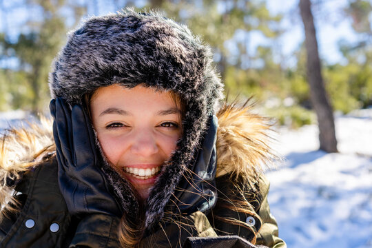 Cheerful woman wearing fur cap smiling while standing on forest during winter