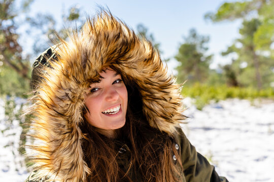 Smiling young woman wearing fur hooded winter coat staring while standing in forest