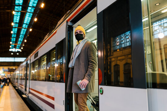 Mid adult man wearing face mask stepping out of train with coffee cup and luggage on station - Powered by Adobe
