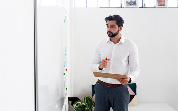 Male entrepreneur with felt tip pen and clipboard looking at whiteboard while working in office