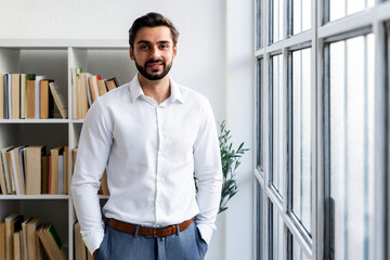 Smiling businessman with hands in pockets standing against bookshelf in office