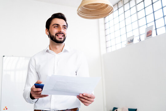 Smiling businessman with paper document on phone call talking through in-ear headphones at work place
