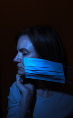 the photo of a frustrated woman with closed eyes in medical mask in a blue light over the black background