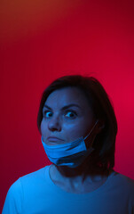 the abstract photo of a frustrated woman with wide opened eyes in medical mask over her chin  in a blue light over the red background
